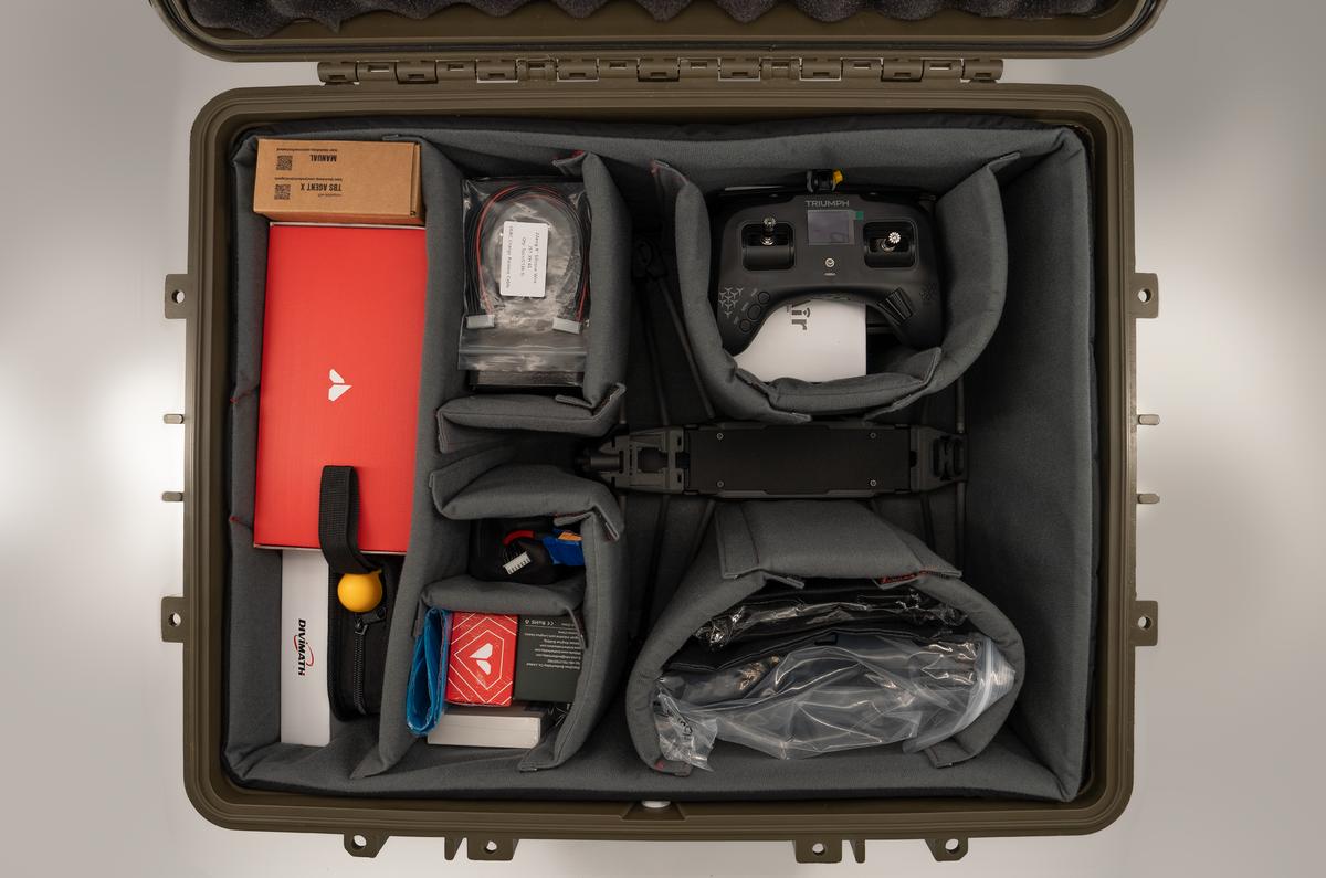 An overhead view of a hard protective carrying case opened wide, filled with drone-related gear in padded compartments. Visible items include a red rectangular device, a black remote controller, a cardboard box, cables, tools, and accessories arranged neatly inside a gray-lined case.