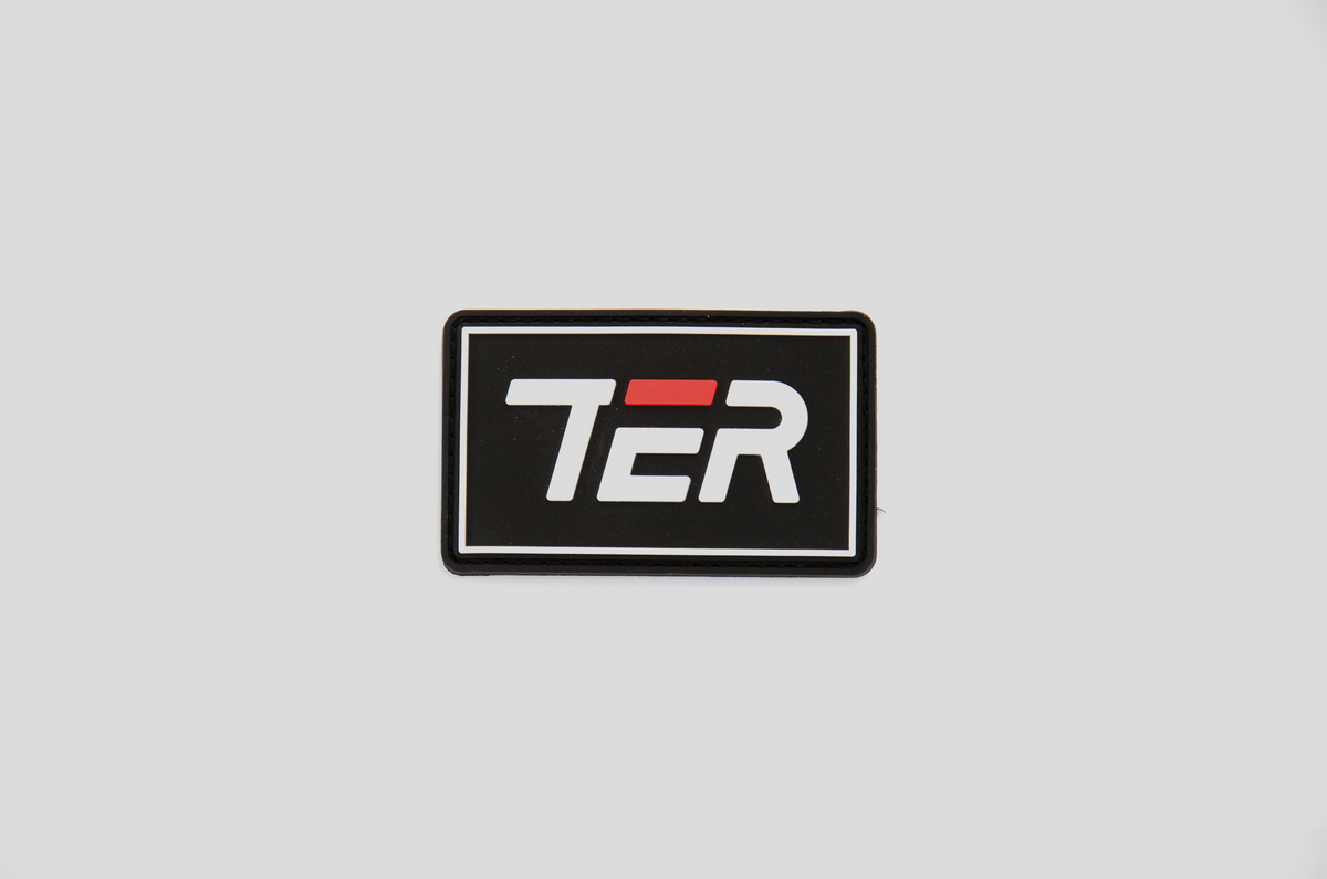 A rectangular black patch with a white border and the stylized letters 'TER' in white, featuring a red accent bar over the middle letter, shown on a light gray background.