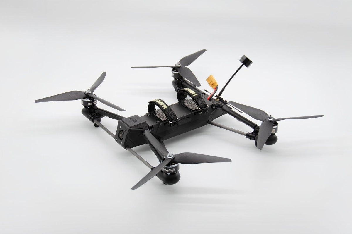A black quadcopter drone with four propellers, a mounted camera, and an antenna, photographed on a clean light gray studio background.
