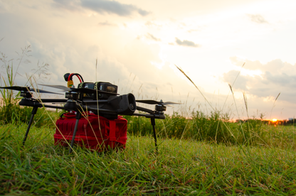 A black quadcopter drone with a red battery case sits in tall green grass outdoors at sunset, with the bright sun and cloudy sky in the background.