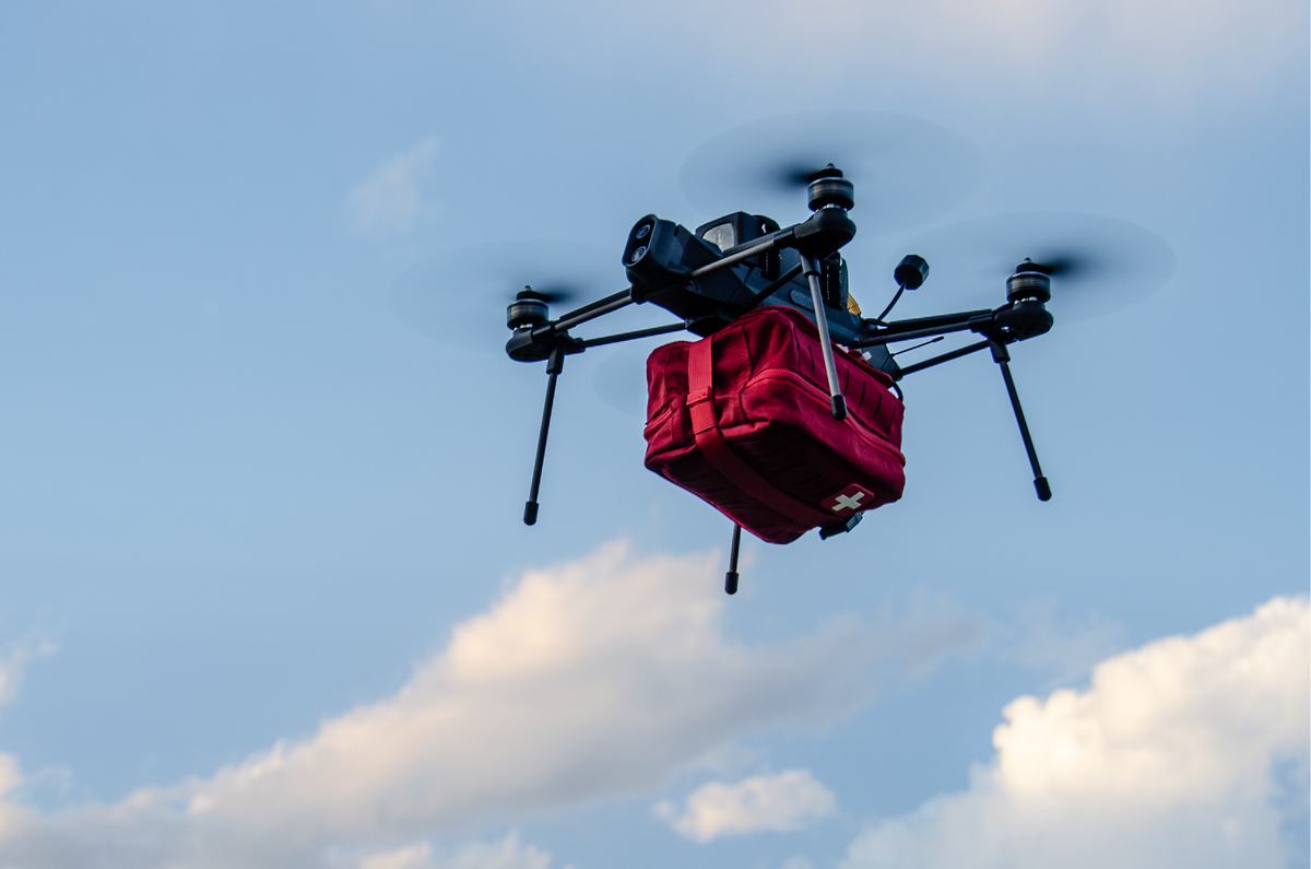 A black quadcopter drone carrying a red insulated delivery bag or package, flying against a blue sky with white clouds.