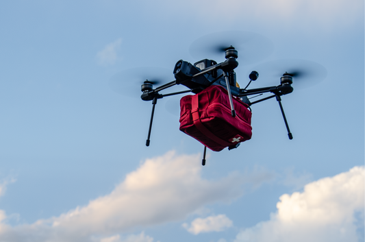 A black quadcopter drone carrying a red insulated delivery bag or package, flying against a blue sky with white clouds.