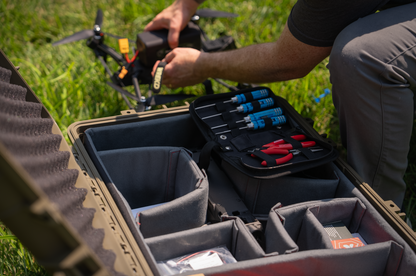 An open hard case on grass containing a drone and a black tool kit with blue-handled screwdrivers and red-handled tools. A person in a black shirt and gray pants is kneeling beside the case, working on the drone in an outdoor grassy setting.
