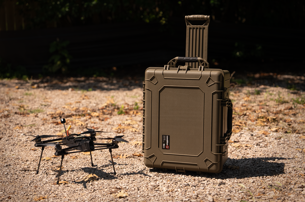 An olive green hard-shell protective case stands on gravel outdoors next to a black quadcopter drone.