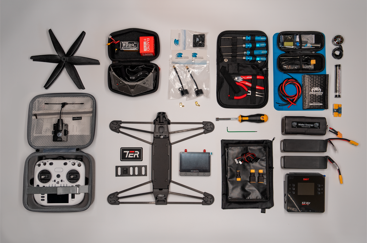 A comprehensive drone kit laid out neatly on a light gray background, including a quadcopter drone, remote controller in an open gray case, spare propellers, batteries, tools in black and blue cases, wiring, and accessories.