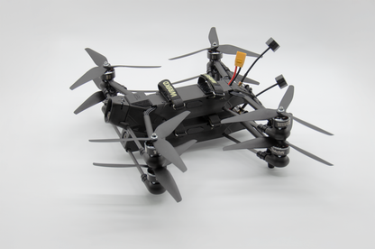 A black quadcopter FPV drone with four gray multi-blade propellers, a visible battery pack, and wiring, photographed on a seamless light gray studio background.