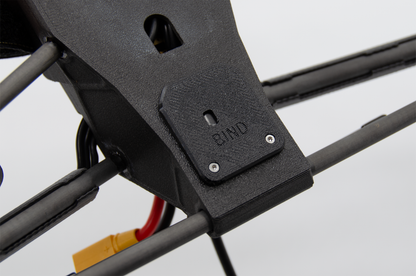 Close-up of a black drone frame component with a small metal screw plate labeled 'BIND' and attached cables, including a yellow and red connector.