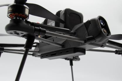 Close-up of a black quadcopter drone with a mounted camera and propellers against a light gray background.