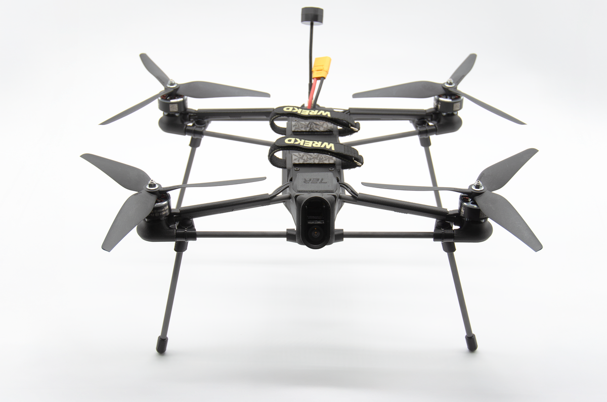 A black quadcopter drone with four propellers and landing legs, photographed from the front against a clean white studio background. The frame has visible yellow and black branding.