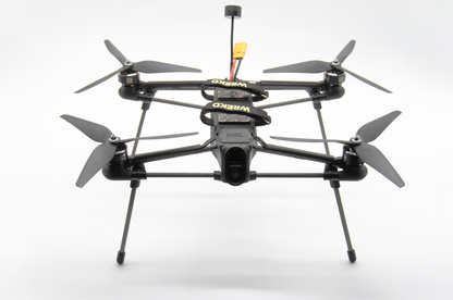 A black quadcopter drone with four propellers and landing legs, photographed from the front against a clean white studio background. The frame has visible yellow and black branding.