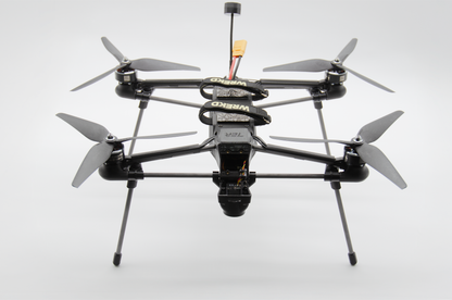 A black quadcopter drone with a mounted camera, four gray propellers, and landing legs, photographed against a seamless light gray studio background.