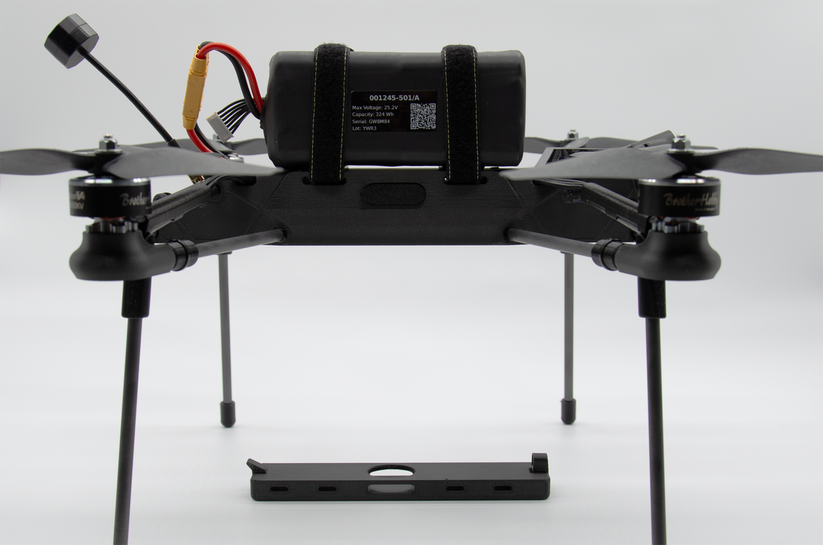 A black quadcopter drone with four propellers, a central battery pack secured by straps, and a separate black mounting plate in front, photographed on a light gray studio background.