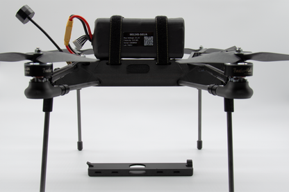 A black quadcopter drone with four propellers, a central battery pack secured by straps, and a separate black mounting plate in front, photographed on a light gray studio background.