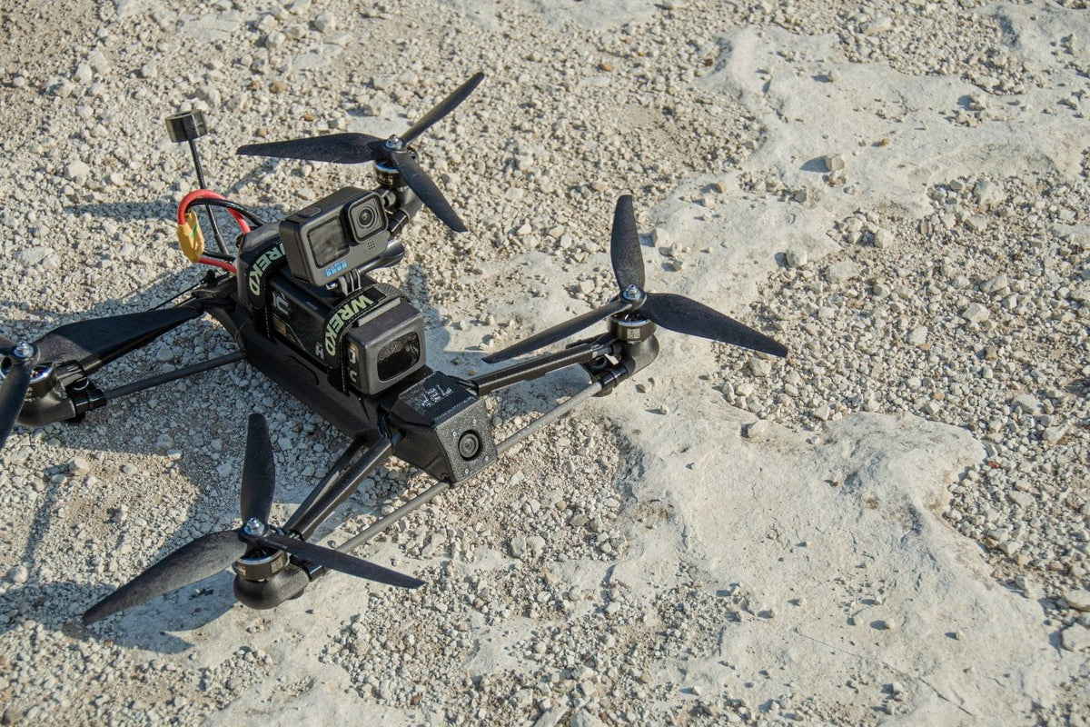 A black quadcopter drone with mounted cameras and propellers, photographed from above on a light sandy, gravel-textured ground.