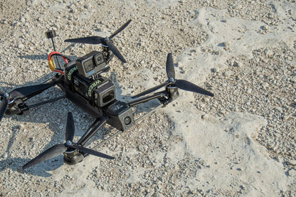 A black quadcopter drone with mounted cameras and propellers, photographed from above on a light sandy, gravel-textured ground.
