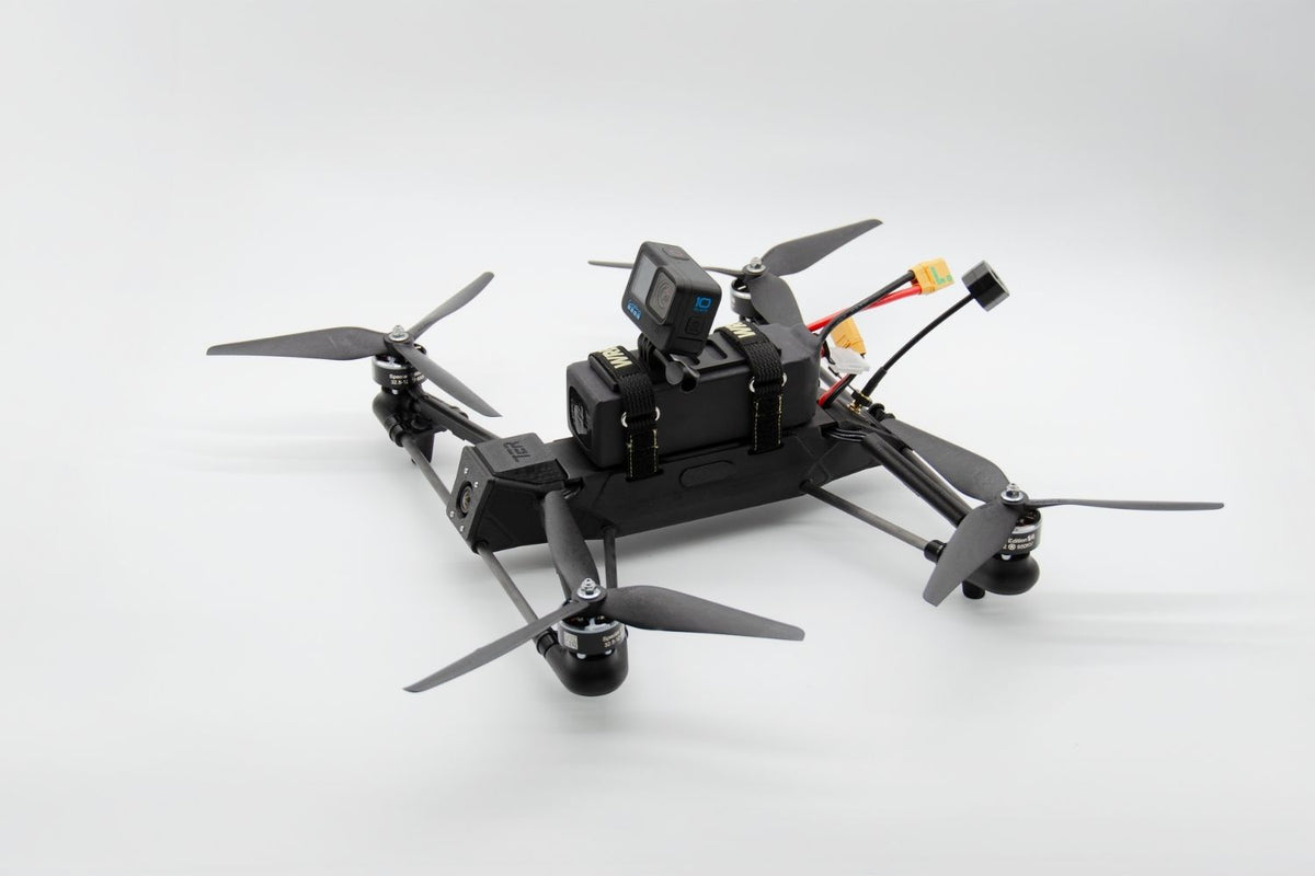 A black quadcopter drone with a mounted action camera on top, shown from a high-angle view against a light gray studio background.