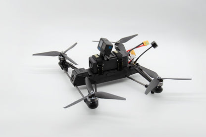 A black quadcopter drone with a mounted action camera on top, shown from a high-angle view against a light gray studio background.