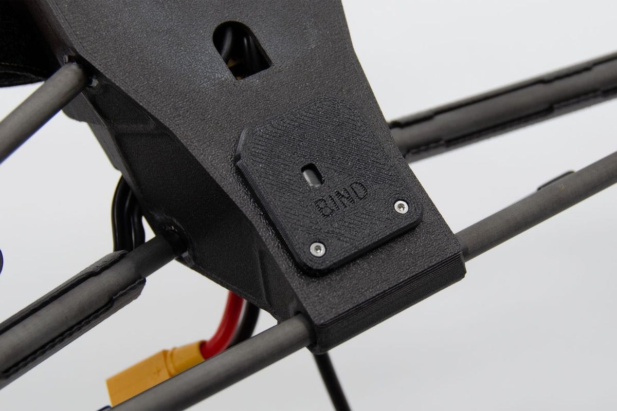 Close-up of a black 3D-printed drone frame component with a small metal screw plate labeled 'BIND' and attached cables, including a yellow and red connector.