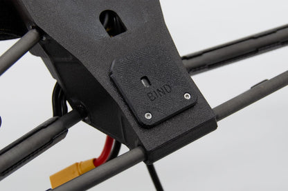 Close-up of a black 3D-printed drone frame component with a small metal screw plate labeled 'BIND' and attached cables, including a yellow and red connector.