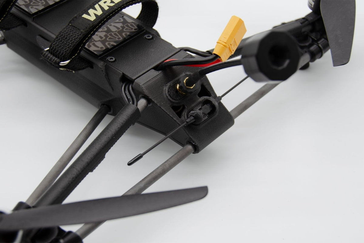 Close-up of a black quadcopter drone body with gray propeller arms, a yellow XT60 connector, and part of a patterned strap on a white background.