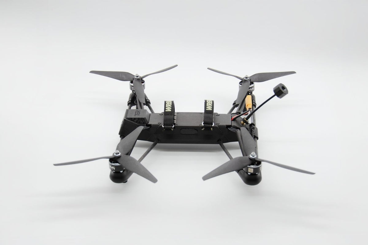 A black quadcopter drone with four gray propellers and a mounted camera, photographed from the rear against a seamless light gray studio background.