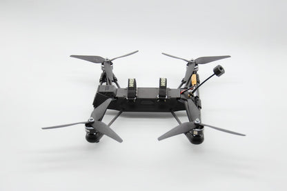 A black quadcopter drone with four gray propellers and a mounted camera, photographed from the rear against a seamless light gray studio background.