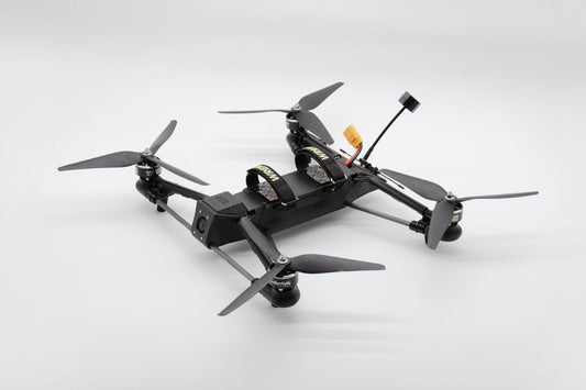 A black quadcopter drone with four propellers, a mounted camera, and an antenna, photographed on a clean light gray studio background.