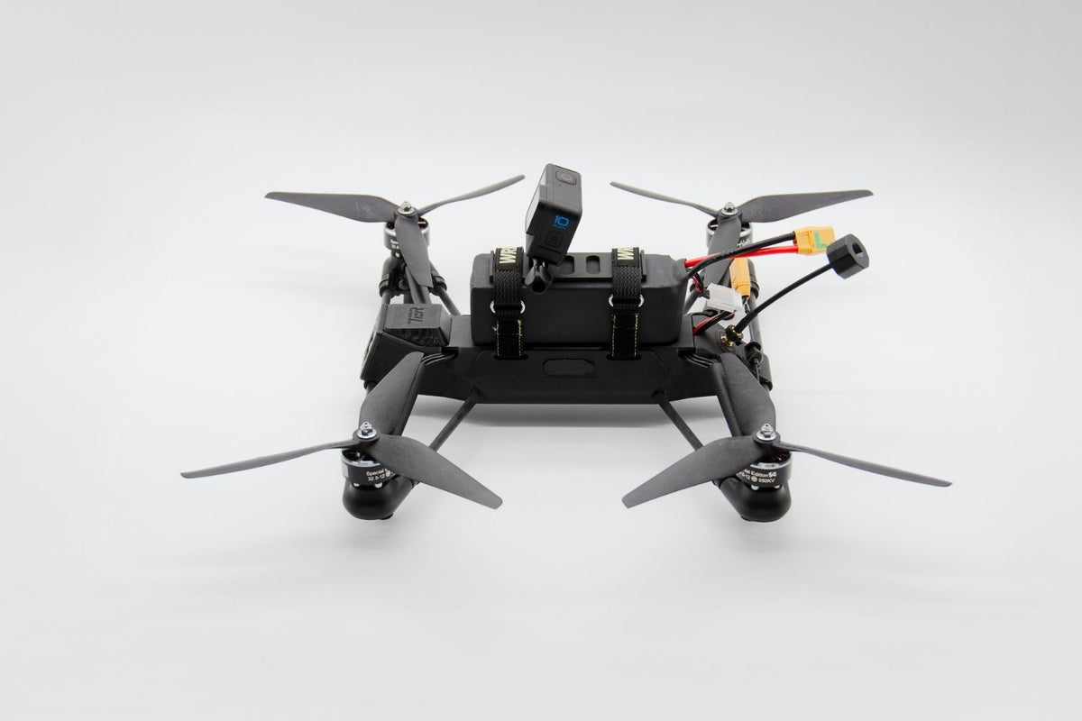 A black quadcopter drone with four gray propellers, a mounted camera, and visible wiring and connectors, photographed on a clean light gray studio background.