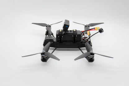A black quadcopter drone with four gray propellers, a mounted camera, and visible wiring and connectors, photographed on a clean light gray studio background.