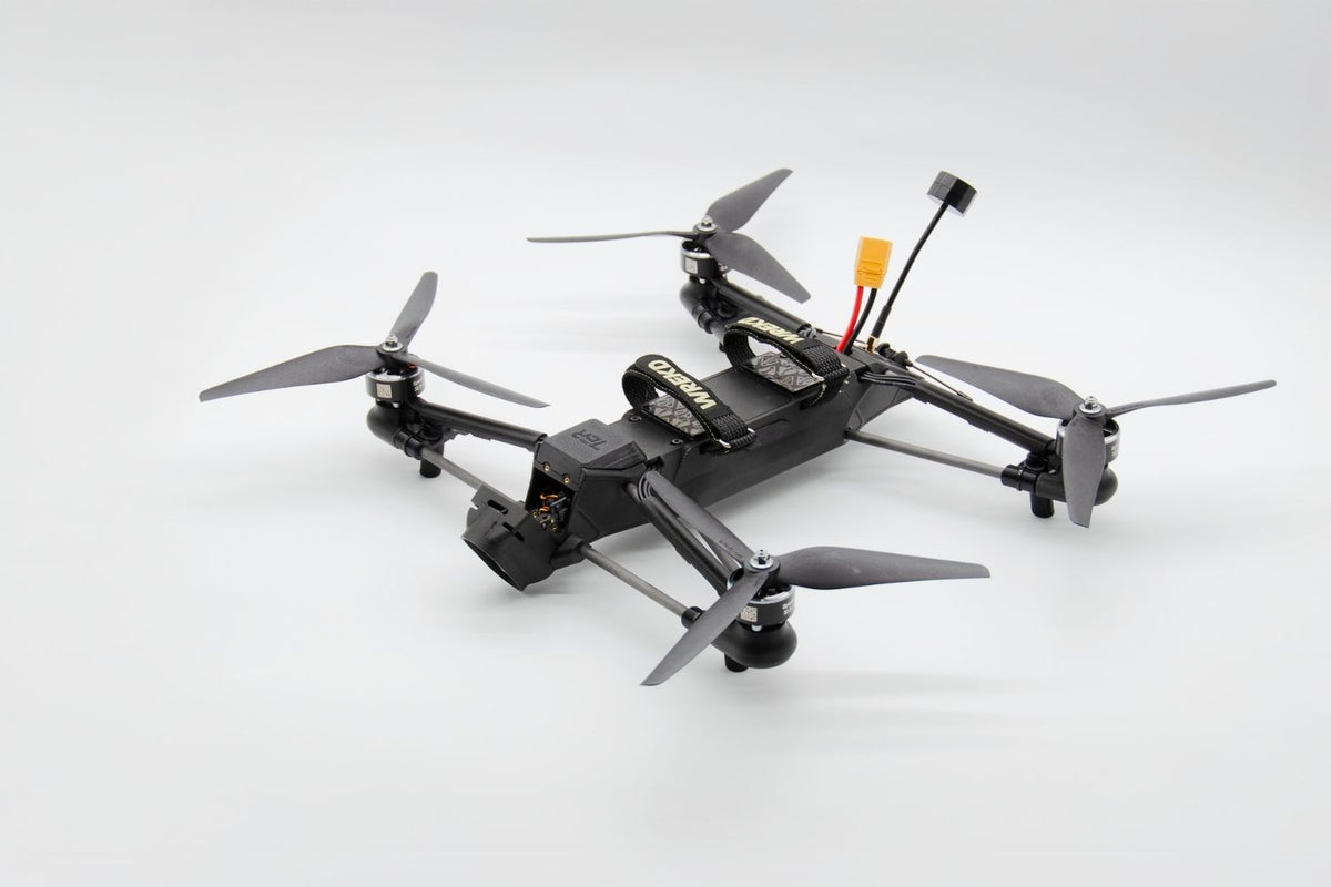 A black quadcopter drone with four gray propellers, a mounted camera, and a small orange connector on a light gray background.