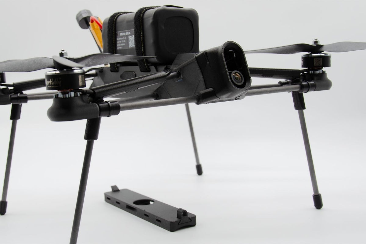 A black quadcopter drone with a mounted battery pack and landing legs, shown on a clean white studio background. A separate black mounting plate is placed in front of the drone.