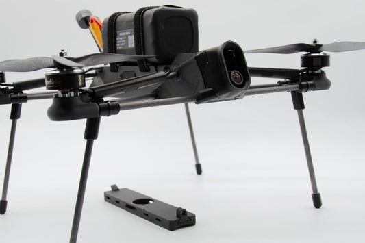 A black quadcopter drone with a mounted battery pack and landing legs, shown on a clean white studio background. A separate black mounting plate is placed in front of the drone.