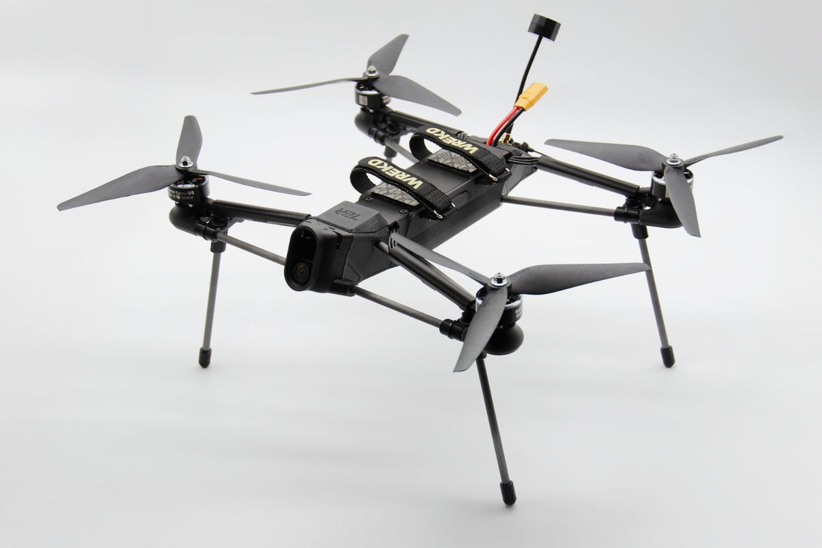 A black quadcopter drone with four propellers, landing legs, and a mounted camera, photographed on a clean light gray studio background.