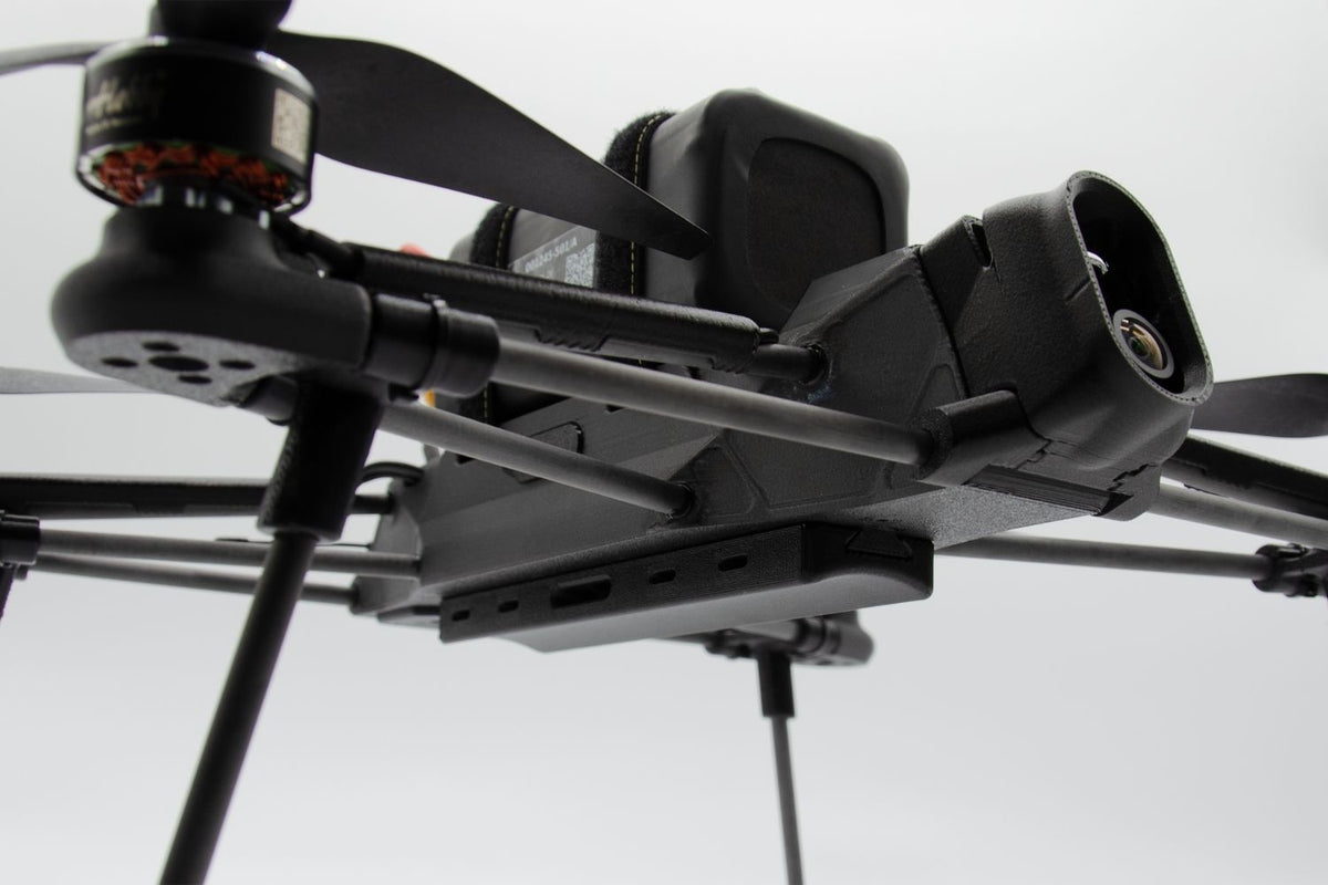 Close-up of a black quadcopter drone with a mounted camera and propellers against a light gray background.