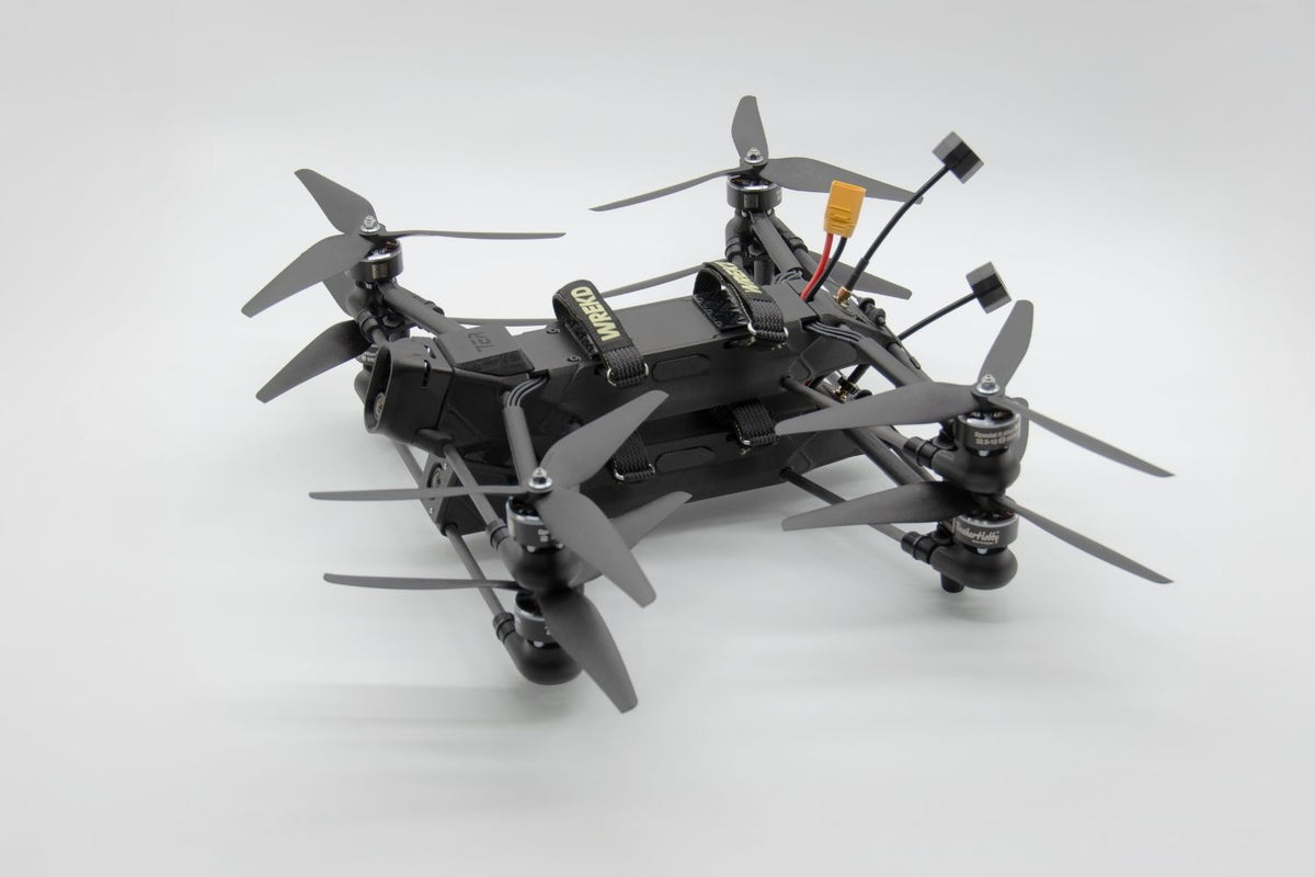 A black quadcopter drone with four gray propellers, a mounted camera, and visible wiring and battery components, photographed on a clean light gray studio background.