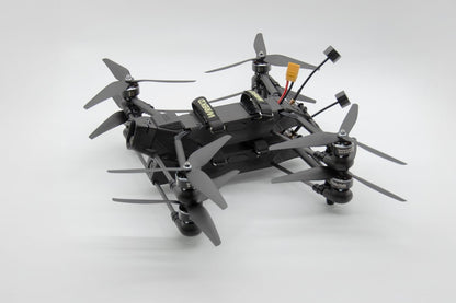 A black quadcopter drone with four gray propellers, a mounted camera, and visible wiring and battery components, photographed on a clean light gray studio background.