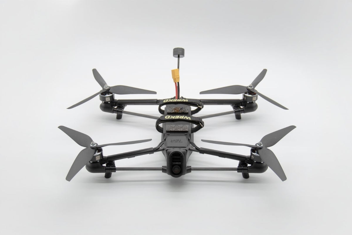 A black quadcopter FPV drone with four propellers and a central battery pack, photographed from a top-down angle on a light gray background.