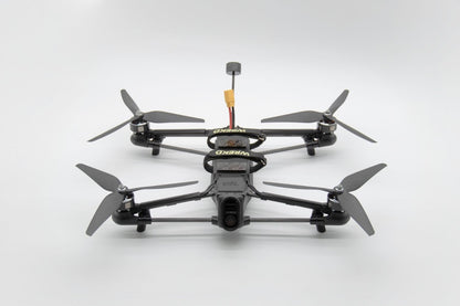 A black quadcopter FPV drone with four propellers and a central battery pack, photographed from a top-down angle on a light gray background.