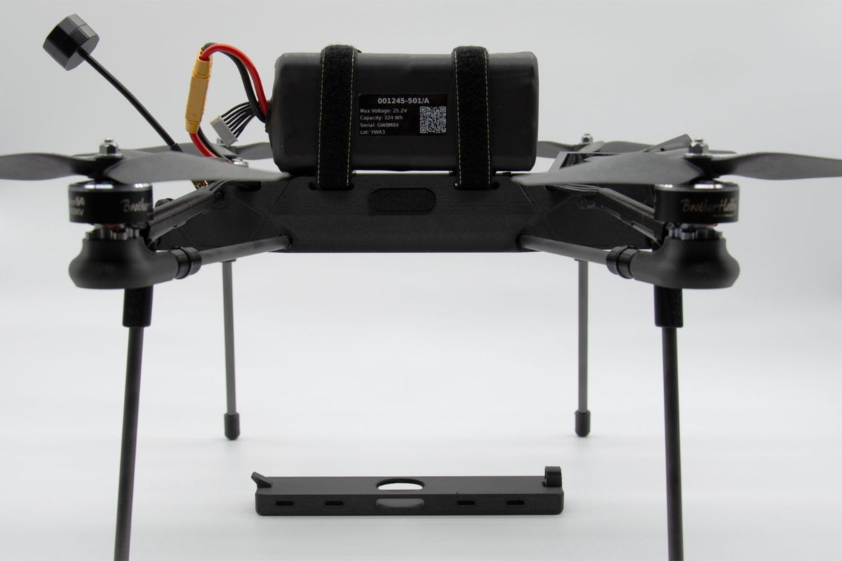 A black quadcopter drone with four propellers, a central battery pack secured by straps, and a separate black mounting plate in front, photographed on a light gray studio background.