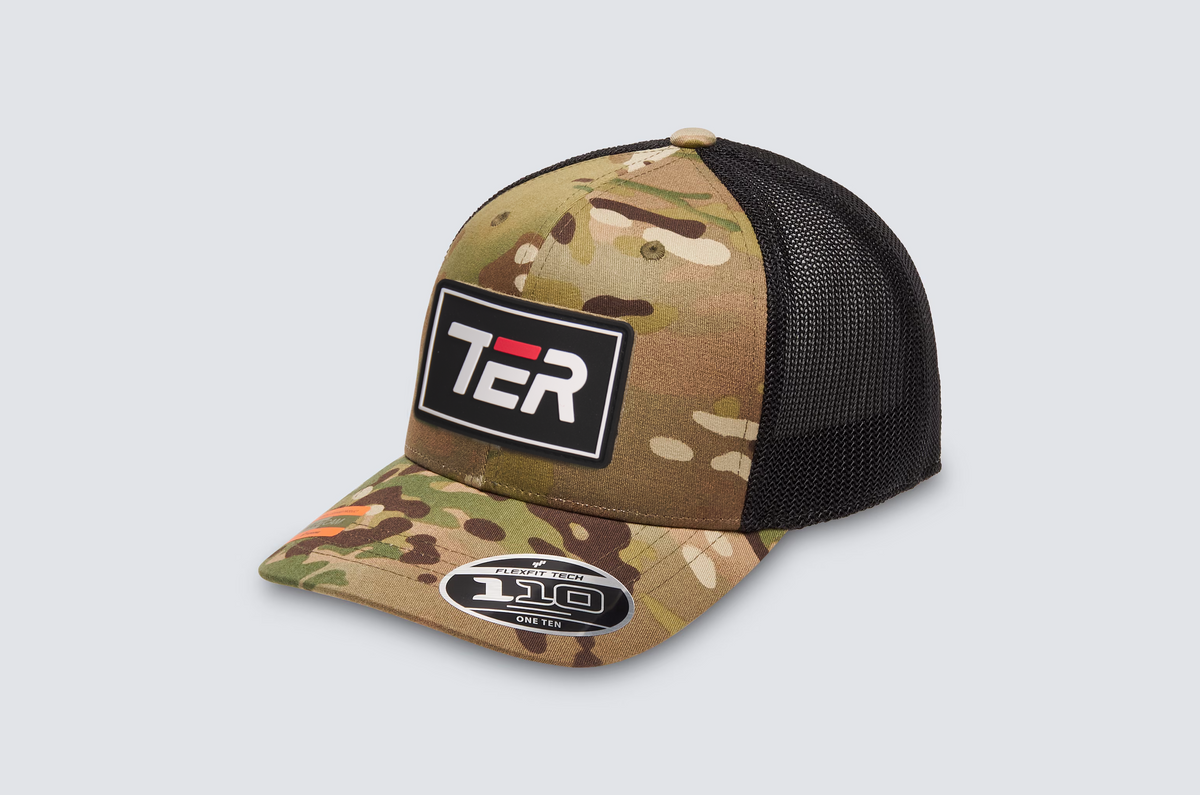 A camouflage trucker hat with a black mesh back and a black rectangular patch on the front displaying 'TER' in white and red. The brim features a sticker for a '110° Flex Fit' cap.