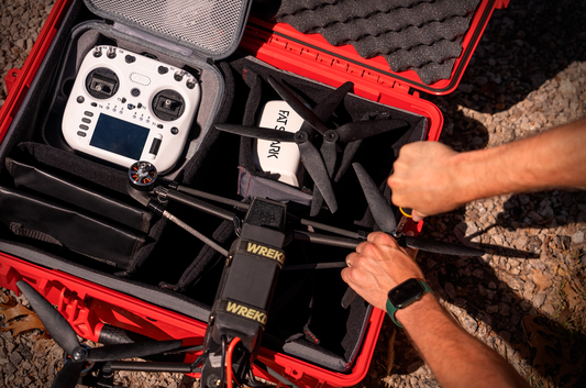 A red hard-shell carrying case opened outdoors on gravel, containing a white FPV drone controller and a black drone with propellers. A person's hands are visible adjusting the drone inside the case.