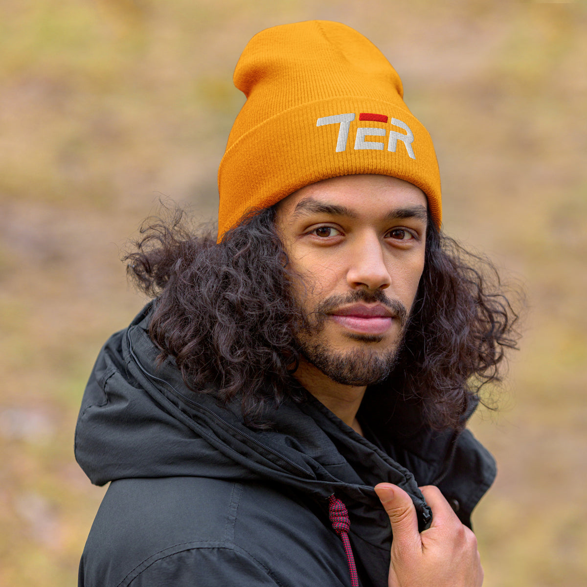 A man with long curly dark hair wearing a gold knit beanie with 'TER' embroidered on the front and a black hooded jacket, photographed outdoors against a blurred natural background.