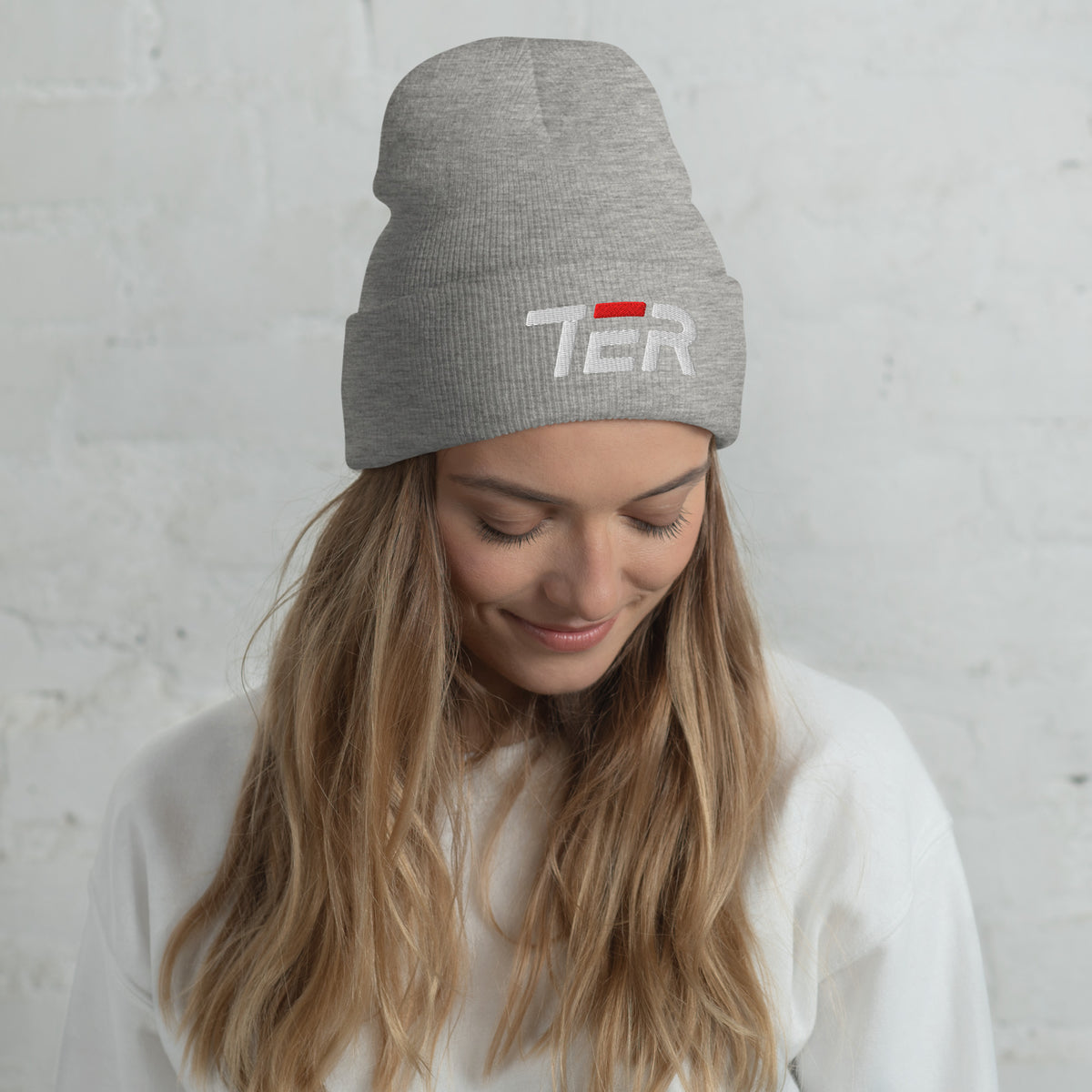 A woman wearing a heather gray knit beanie with a white embroidered 'TER' logo and a small red accent, photographed against a white brick wall.