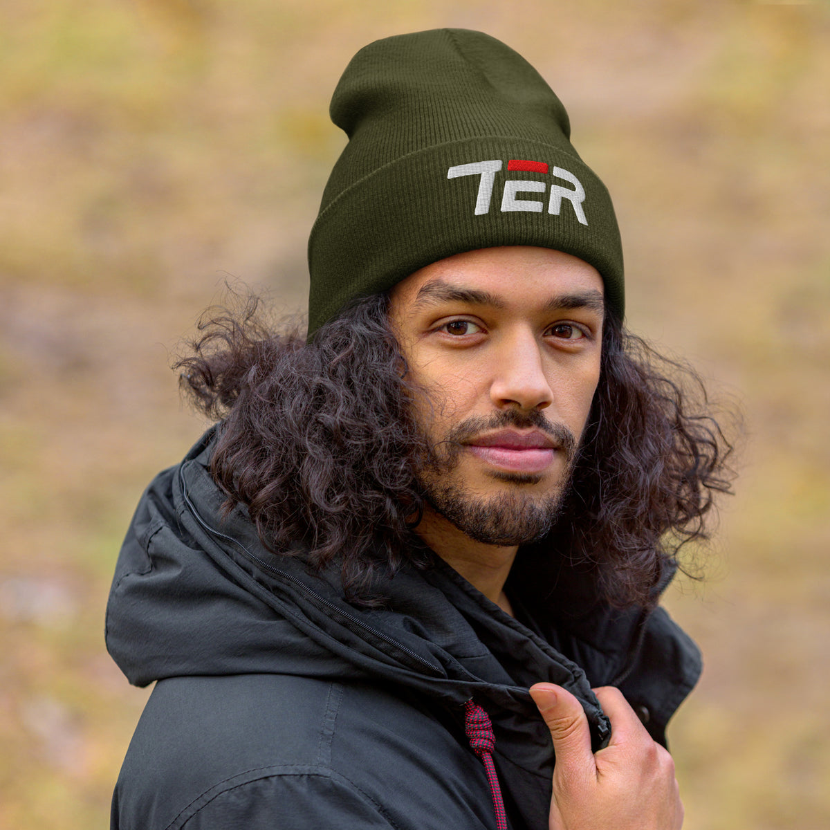 A man wearing an olive green knit beanie with 'TER' embroidered in white on the front, paired with a black hooded jacket, set against a blurred natural background.
