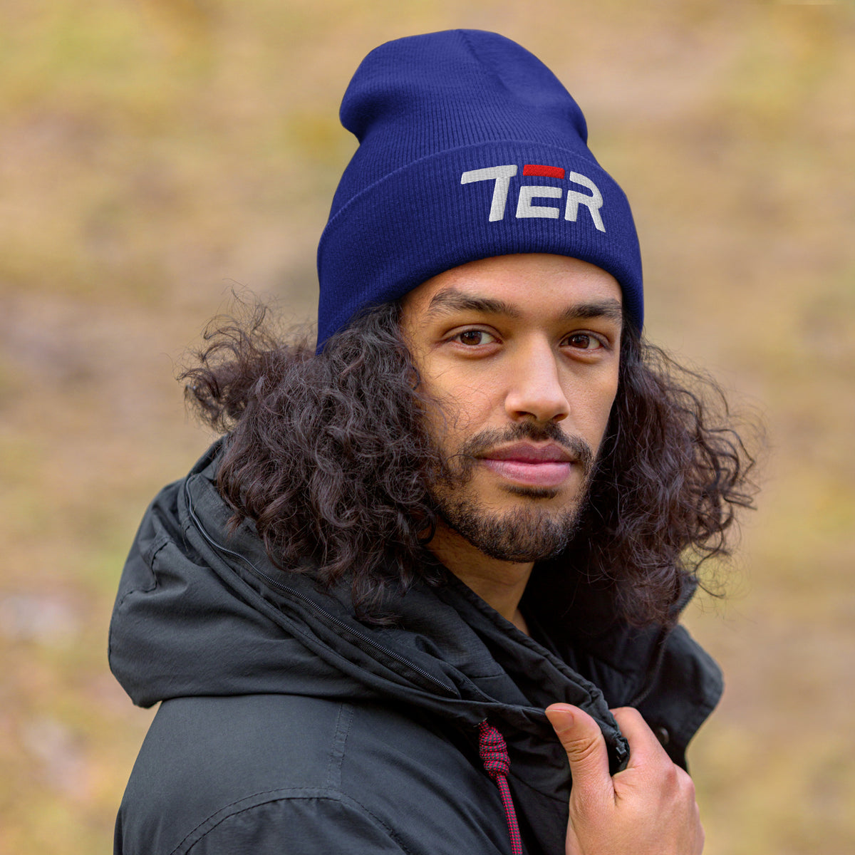 Man wearing a blue beanie with text and a logo, standing outdoors with a blurred natural background.