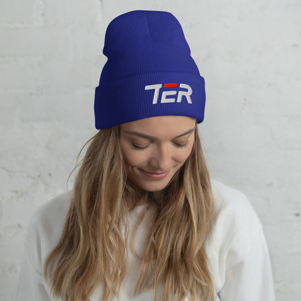 A woman wearing a royal blue ribbed knit beanie with a white and red embroidered 'TER' logo on the front, photographed against a white brick wall.